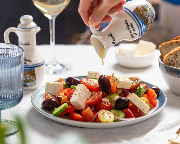 A Greek salad is on a table. Olive oil is being poured on it.