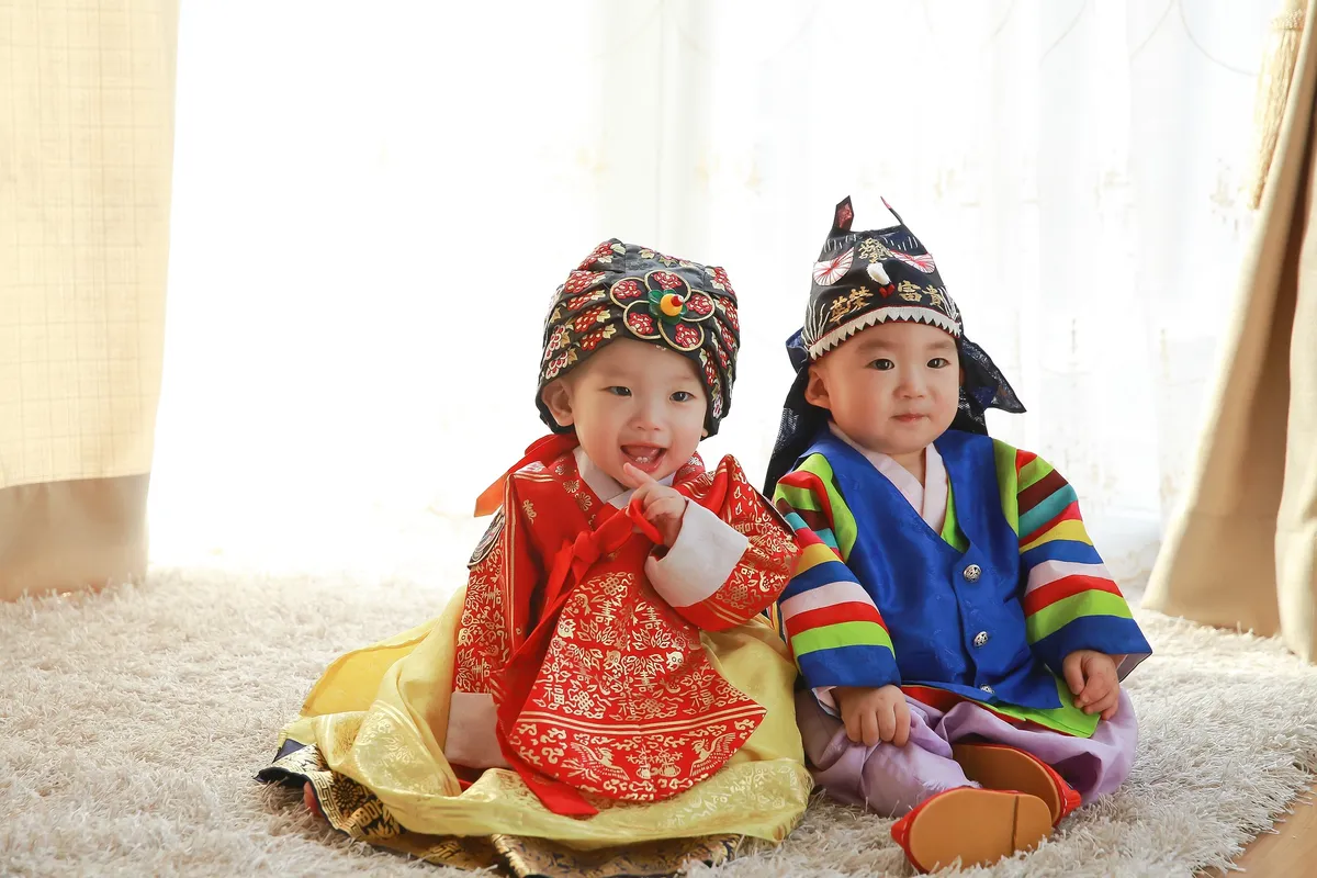 Two babies in traditional Korean clothing. They are sitting on white rug, and there is a light coming through the curtain behind them.