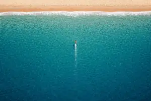 Landscape aerial drone view of a tiny lone swimmer in the open ocean heading straight toward a sandy beach at the top of the frame, leaving a narrow wake behind across a vast expanse of blue water.