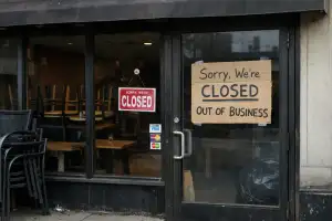A street-level photo of a recently closed restaurant with dark windows, stacked chairs inside, a closed sign, and a handmade cardboard “out of business” notice taped to the glass door.