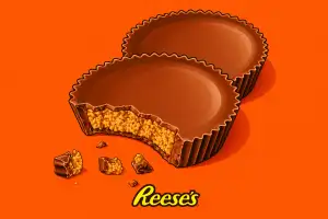 Illustrated landscape image of two Reese’s peanut butter cups on a bright orange background, with one cup bitten open to show the peanut butter filling and small chocolate crumbs below.