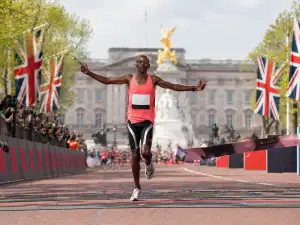 A marathon runner in a coral top and black shorts crosses a London finish line with arms outstretched, with Union Jack flags, spectators, and Buckingham Palace in the background.