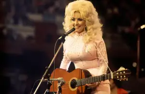A woman with large blond hair smiles while performing on stage with a guitar and microphone.