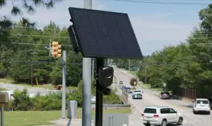 A pole-mounted roadside camera beneath a large solar panel overlooks a street with several passing cars.