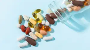 A clear bottle tipped on its side spills assorted pills and soft-gel capsules onto a light blue surface.