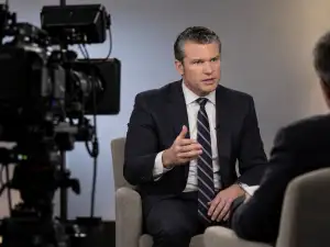 A man in a dark suit gestures while speaking to another seated person, with a large television camera in the foreground.