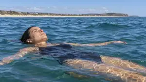 A swimmer in a dark swimsuit floats on their back in the sea, arms spread and face above water, with a sandy coastline in the distance.
